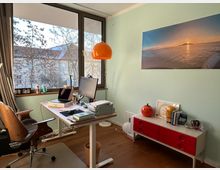 Helles Arbeitszimmer mit einem großen Fenster, durch das Bäume und Nachbarhäuser sichtbar sind. Ein Schreibtisch mit Computer, Büromaterialien und einem Stuhl steht vor der Fensterfront; an der mintgrünen Wand hängt ein Bild mit Sonnenuntergangsmotiv, daneben eine rote Kommode mit Dekorationsgegenständen und Büchern.