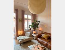 Wohnzimmer mit gemütlicher Einrichtung: ein braunes Ledersofa mit Kissen, ein helles Polster-Liegeelement mit Felldecke, ein marmorierter Couchtisch mit Kerzen und Geschirr sowie eine große Zimmerpflanze neben einem Heizkörper unter hohen Doppelfenstern mit beige Vorhängen. Eine große, runde Papierlampe hängt von der Decke, und kleine Kerzen sind im Raum verteilt.