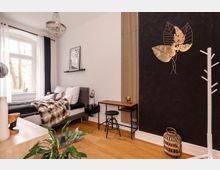 Schlafzimmer mit modernem Design, ausgestattet mit einem grauen Polsterbett, angrenzendem kleinen Holztisch mit schwarzem Hocker und schwarzen Nachttischen. Die Wände sind teils weiß und teils mit schwarzer Tapete sowie vertikalen Holzleisten gestaltet, dekoriert mit einer goldfarbenen Blätterwandverzierung. Ein großes Fenster mit weißen und dunklen Vorhängen sorgt für Tageslicht, während eine kristallene Deckenleuchte und Holzboden das Ambiente abrunden.