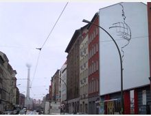 Straßenansicht in einer Stadt mit mehrstöckigen Gebäuden, teilweise mit Fassadenkunst. Im Hintergrund ist der Berliner Fernsehturm sichtbar, dessen Spitze im Nebel verschwindet. Straßenlaternen und Oberleitungen für Straßenbahnen verlaufen entlang der Straße.