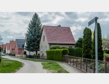 Eine ruhige Wohnstraße in einem Vorort mit freistehenden Einfamilienhäusern, jeweils mit roten oder dunklen Dachziegeln. Vorgärten mit Hecken, Bäumen und Blumenbeeten säumen den Gehweg, und der Himmel ist bewölkt.