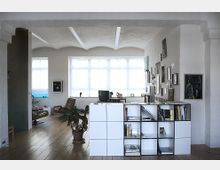 Helles Wohnzimmer oder Arbeitszimmer in einem Loft mit weiß verputzten Wänden und gewölbter Decke. Der Raum ist mit Holzdielenboden, einem Regal aus weißen Würfeln, Zimmerpflanzen, Bildern an den Wänden und großen Fenstern mit viel Tageslicht gestaltet.