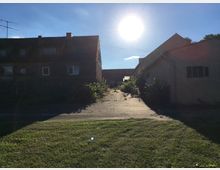 Eine ländliche Szenerie mit zwei alten Gebäuden aus Stein und Putz, die einen schmalen, überwucherten Weg säumen. Die Sonne steht tief am Himmel, beleuchtet das Gelände und wirft lange Schatten, während im Vordergrund eine Grasfläche zu sehen ist.