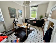 Arbeitszimmer mit einem Holztisch in der Mitte, zwei Monitoren und Kabelmanagement-Armen. Der Raum hat cremefarbene Wände, ein großes Fenster mit Blick auf grüne Bäume, eine niedrige schwarze Schrankwand mit Büchern und Dekoration, sowie einen rot-weißen Fliesenboden und eine helle Deckenleuchte.