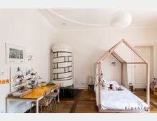 Helles Kinderzimmer mit einem weißen Kachelofen, einem Schreibtisch mit Bastelmaterialien auf der linken Seite und einem Bett in Form eines Holzhausgestells rechts. Der Boden besteht aus Parkett, und die Wände sind schlicht weiß gehalten.