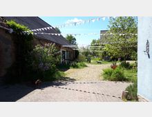 Eine ländliche Hofanlage mit Kopfsteinpflaster und umliegenden Gebäuden aus Backstein. Über den Hof spannen sich weiße Wimpelgirlanden, Bäume und Sträucher spenden Schatten, und in der Mitte stehen Sitzgelegenheiten und Pflanzen in Töpfen.