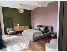 Modern bedroom with a low upholstered bed, green accent wall and pink side wall, and herringbone wood flooring. The room includes pendant lights, a wooden sideboard, an armchair, a round shag rug, and a small green side table.