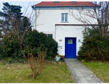 Zweistöckiges weißes Wohnhaus mit rotem Satteldach, einem blauen Holzeingang und einem gepflasterten Gehweg, umgeben von Sträuchern, Bäumen und Rasen.