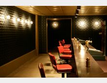 Barraum mit Theke und roten Barhockern, beleuchtet durch Wandlampen und Deckenleuchten. Die Wände sind dunkel und weisen ein strukturiertes, gemustertes Design auf.