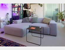 Modernes Wohnzimmer mit großem hellgrauem Ecksofa, kleinem schwarzem Metall-Couchtisch und mehreren Zimmerpflanzen. Im Hintergrund stehen offene Regale und eine Kleiderstange, daneben eine Treppe sowie eine Milchglas-Schiebetür.