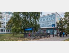 Außenansicht des U-Bahnhofs Amrumer Straße in Berlin mit einem blau-weiß gemusterten Gebäude im Hintergrund, Fahrradständern im Vordergrund und mehreren Personen, die sich vor dem Eingang aufhalten. Bäume und Büsche umgeben die Szene.