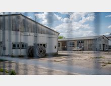 Industriegelände mit verfallenen Hallen aus Beton, verblasster Fassade und heruntergekommenen Fenstern. Im Vordergrund stehen Kabeltrommeln, im Hintergrund ist ein älteres weißes Auto vor einem verschlossenen Tor zu sehen.