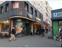 Straßenszene mit einem Eckgebäude, das mehrere Geschäfte beherbergt, darunter eine Bäckerei mit der Aufschrift „La Femme“, ein Café namens „Lookma“ und eine Apotheke. Rechts im Bild befindet sich der Eingang zur U-Bahn-Station „Bismarckstraße“ mit einem grünen Kachelmuster und einem blauen „U“-Schild.