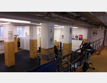Ein heller, rechteckiger Fitnessraum mit gelb-weißen Säulen, einem Boxsack, Trainingsgeräten und Postern an den Wänden. Im Vordergrund befindet sich eine Tribüne mit Sitzbank, im Hintergrund eine Trainingsfläche mit Seilen abgetrennt.