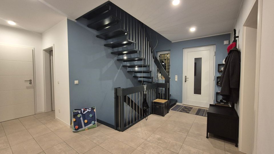 Moderne Eingangshalle in einem Wohnhaus mit einer schwarz-grauen freitragenden Treppe an einer blauen Wand. Die Halle hat helle Fliesenböden, eine weiße Eingangstür mit eingelassener Glasscheibe und eine Garderobe mit Haken und Ablagen an der rechten Seite.