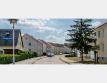 Wohnstraße mit Einfamilienhäusern und Reihenhäusern, teilweise mit rot gedeckten Dächern und hellen Fassaden. Die Straße ist von gepflegten Hecken gesäumt, mit einem Gehweg und Laternen; ein großer Nadelbaum steht auf einer begrünten Fläche am Straßenrand.