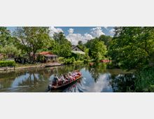 Eine Wasserlandschaft mit einem ruhigen Fluss, umgeben von dichtem Grün und einzelnen Häusern mit roten Dächern am Ufer. Im Vordergrund fährt ein Boot mit mehreren sitzenden Personen, während im Hintergrund kleine Kanus auf dem Wasser treiben.