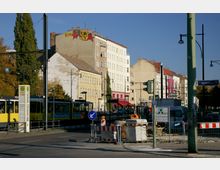 Straßenkreuzung in einer Stadt mit mehrstöckigen Wohnhäusern, einer gelben Straßenbahn an einer Haltestelle und Baustellenabsperrungen im Vordergrund. Straßenlaternen und Schilder umgeben den Bereich, während im Hintergrund Bäume und klare Herbstsonne zu sehen sind.