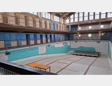 Ehemaliges Schwimmbad mit leeren, trockenen Becken, türkisfarbenen Fliesen und teils abgebröckelten Wänden. Im oberen Bereich sind Umkleidekabinen und Geländer sichtbar, begleitet von großen Fenstern, die natürliches Licht hereinlassen.