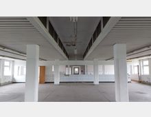 Ein leerer, rechteckiger Raum mit weißen Wänden, Fliesen an der unteren Hälfte der Wände und mehreren stabilen weißen Säulen. Die Decke hat Lüftungsschlitze und sichtbare Gebrauchsspuren, während mehrere Fenster Tageslicht einlassen.