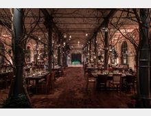 Großer Saal mit hohen Decken und dekorativen, baumähnlichen Strukturen, die von metallischen Säulen getragen werden. Der Raum ist mit reich gedeckten Tischen, hängenden Laternen und kleinen Lichtern beleuchtet, während der Holzboden mit herbstlichem Laub bedeckt ist.