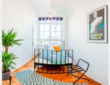 Helles Schlafzimmer mit einem schmalen Bett mit schwarzem Metallrahmen, weißen und grünen Betttextilien sowie einem gelben Kissen. Der Raum hat Holzboden, eine große Fensterfront mit gestreifter Gardine, einem Zimmerpflanze in einer Ecke und einem gerahmten Tierpark-Berlin-Poster an der Wand.