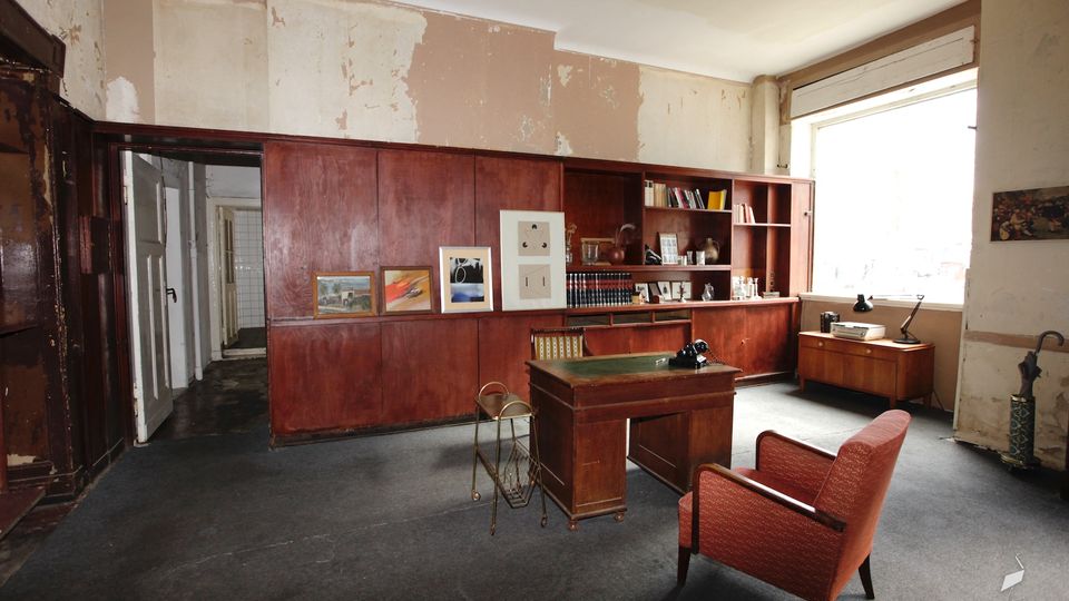 Geräumiges Büro mit großer dunkler Holzschrankwand und offenen Regalen, davor ein Holzschreibtisch und ein roter Polstersessel auf grauem Teppichboden. Helles Fenster an der rechten Seite, abblätternder Putz an den Wänden und eine Türöffnung links zu einem Flur.