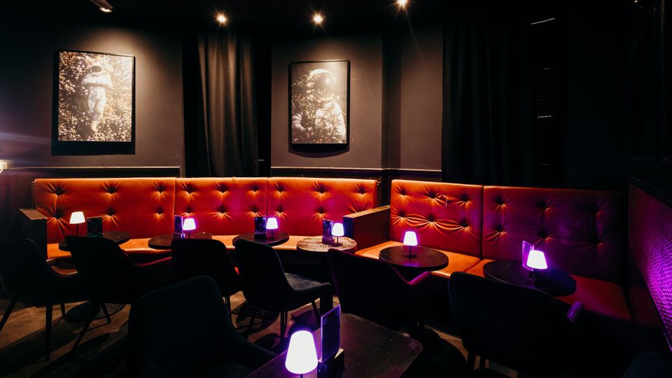 Gedimmter Lounge- oder Barbereich mit orangefarbenen, tiefgepolsterten Sitzbänken entlang der Wände, kleinen runden Tischen und schwarzen Sesseln. Dunkle Wände mit Vorhängen, Spotbeleuchtung an der Decke, lilafarbenen Tischlampen und gerahmten Bildern an der Wand.
