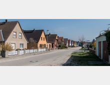 Wohnsiedlung mit Einfamilienhäusern in einer ruhigen Straße, geprägt von verschiedenen Fassadenfarben und Satteldächern. Weiße und rote Zäune sowie kleine Vorgärten trennen die Häuser von der Straße.