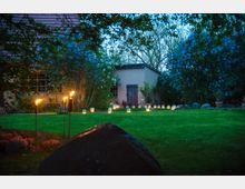 Ein beleuchteter Garten am Abend mit lila blühenden Büschen, einer Rasenfläche und einer hell beleuchteten Reihe von Laternen. Im Hintergrund steht ein helles Gebäude mit Holzfenster und -tür, umgeben von dichter Vegetation.