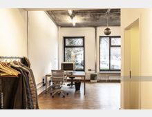 Helles Büro mit Parkettboden, einer Garderobe links, einem Schreibtisch mit Stuhl und Computer in der Mitte sowie großen Fenstern, die Blick auf eine Baumreihe und eine Straße bieten. Die Decke besteht aus sichtbar belassenem Beton mit einfacher Beleuchtung.