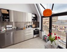 Moderne Küche mit Edelstahlfronten, Einbauspüle, Geschirrspüler und Backofen. Große Fensterfront mit Blick auf städtische Gebäude, ergänzt durch eine orangefarbene Hängeleuchte und eine Vase mit roten Tulpen auf einem kleinen Tisch.