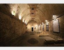 Historischer Gewölbekeller mit Backsteinwänden und leicht gewölbter Decke, beleuchtet durch Wandlampen. An den Wänden hängen Schautafeln, auf dem Boden sind Ausgrabungen und bauliche Reste sichtbar.