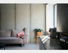 Modernes Wohn- und Kücheninterieur mit Sichtbetonwand, grauem Sofa und Teppich sowie einer Topfpflanze auf einem kleinen Beistelltisch. Rechts fällt Tageslicht durch ein großes Fenster auf eine Küchenarbeitsplatte mit Marmoroptik und mehrere Küchengeräte.