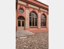 Historische Gebäudefassade in Rosé mit großen Rundbogenfenstern und dekorativen Stuckelementen zwischen hellen Ziegelpfeilern. Links führt eine kurze Steintreppe zu einer zweiflügeligen Eingangstür, davor liegt ein gepflasterter Innenhof aus Kopfsteinpflaster.