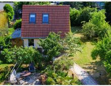 Ein kleines Einfamilienhaus mit rotem Ziegeldach und zwei Dachfenstern, umgeben von einem grünen Garten mit Bäumen, Büschen und einem Steinpfad. Im Vordergrund befinden sich Gartenmöbel und eine gemütliche Sitzgelegenheit auf einer gepflasterten Terrasse.