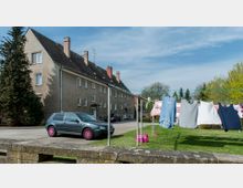 Mehrfamilienhaus mit grauer Fassade und steilem Satteldach, im Vordergrund eine Wiese mit gespannter Wäscheleine und aufgehängter Kleidung. Ein graues Auto mit pinken Felgen steht auf der gepflasterten Fläche links vor dem Gebäude, umgeben von Bäumen und blauem Himmel.