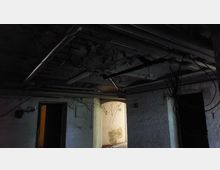 Ein dunkler, heruntergekommener Kellerraum mit weißen gemauerten Wänden und stark beschädigter Decke, an der Rohre und Kabel sichtbar sind. Ein beleuchteter Durchgang und ein Notausgangsschild sind zu erkennen.