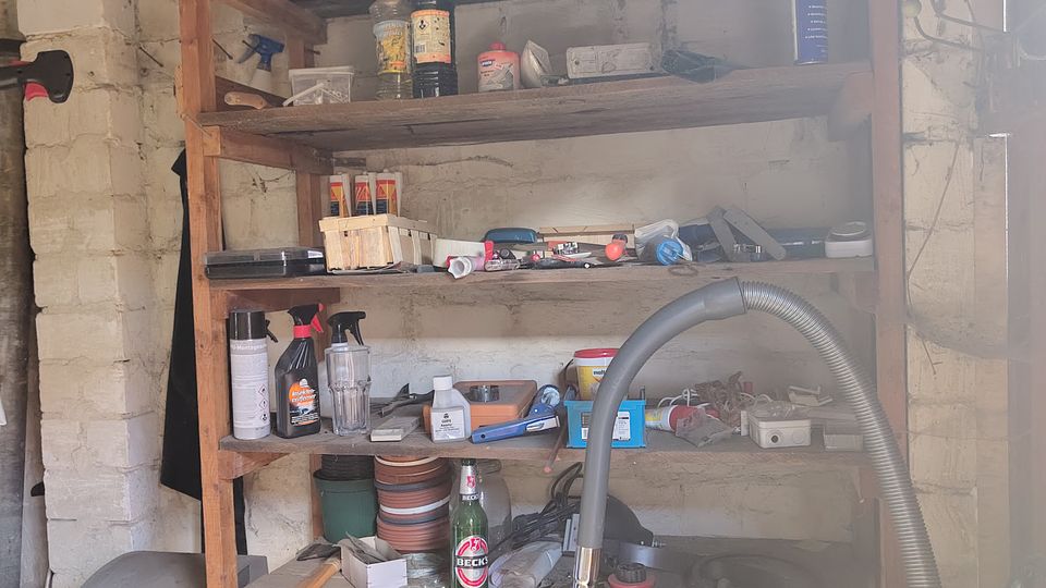 Innenansicht eines kleinen Abstellraums oder einer Werkstatt mit weißen Ziegelwänden und einem großen Holzregal voller Werkzeuge, Reinigungsmittel und verschiedener Gegenstände. Im Vordergrund stehen ein Staubsaugerschlauch und weitere gelagerte Utensilien, darüber verläuft eine Deckenleuchte unter einer Holzbalkendecke.