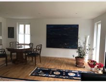 Wohnzimmer mit Holzboden und einem runden Holztisch, umgeben von vier Stühlen. An der weißen Wand hängt ein großes, dunkles Kunstgemälde, daneben steht eine Pflanze in einem Terrakottatopf. Ein Teppich mit blauem Muster bedeckt einen Teil des Bodens, und auf der rechten Seite sind Fenster mit Tageslicht sowie ein paar Blumentöpfe sichtbar.