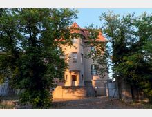 Dreistöckiges historisches Haus mit hellbrauner Fassade und einem roten Walmdach, umgeben von hohen Bäumen. Der Eingang des Gebäudes ist durch eine kleine Treppe zugänglich und von ovalen Fenstern flankiert.