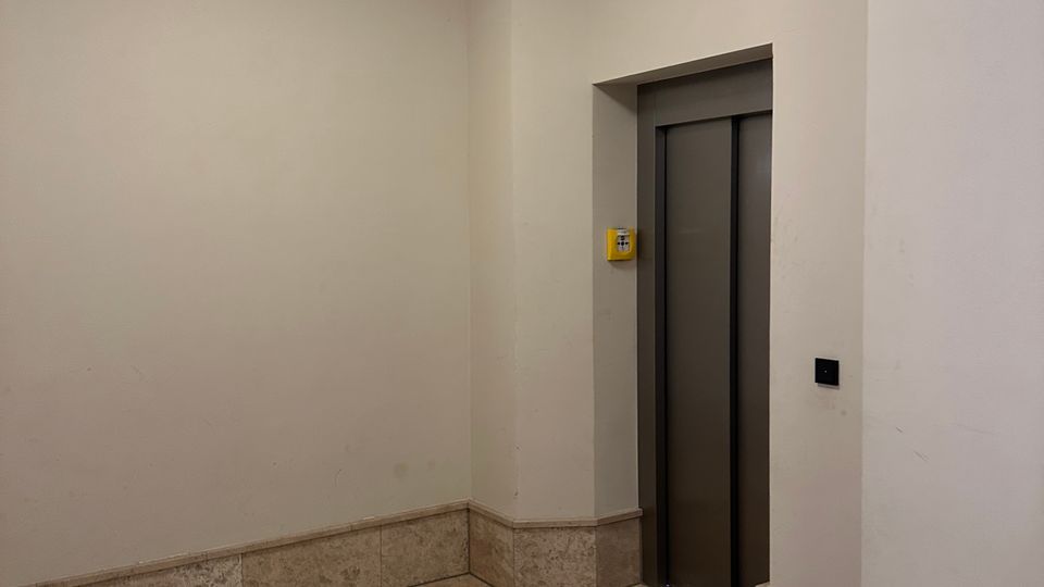 Kleines, helles Treppenhaus- oder Flurfoyer mit beigen Wänden, Steinfliesenboden und einer quadratischen Deckenleuchte. Rechts befindet sich eine eingelassene Aufzugstür mit Bedientaster an der Wand.