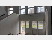 Treppenhaus mit weiß verputzten Wänden und mehreren Fenstern, die nach draußen auf eine Straße und grüne Wiese blicken; im Erdgeschoss zwei Glastüren, von denen die linke offen steht.