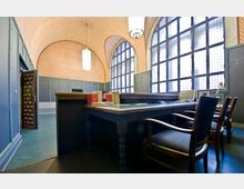 Innenraum eines historischen Büros mit hohen, gewölbten Decken und einem geometrischen Ziegelmuster. Große Gitterfenster lassen Tageslicht herein, während ein massiver Schreibtisch mit Büchern, einem Computer und Lampen im Vordergrund steht; links ist ein offener Tresorraum erkennbar.