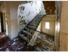 Innenansicht eines stark verfallenen Treppenhauses in einem alten Gebäude mit abblätternder Farbe an den Wänden, bröckelndem Putz und Schutt auf den Stufen. Eine zweiläufige Treppe mit Metallgeländer führt um ein Treppenauge, daneben fällt Licht durch ein kleines Fenster auf das Podest.