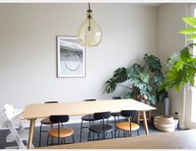 Essbereich mit einem hellen Holztisch, umgeben von schwarzen und orangenen Stühlen sowie einem weißen Kinderstuhl. Im Hintergrund stehen große grüne Pflanzen, ein Korb und ein gerahmtes Schwarz-Weiß-Bild an der beigen Wand, während eine große Glühbirnen-Pendelleuchte von der Decke hängt.