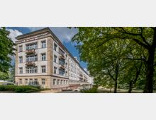 Ein mehrstöckiges Wohn- oder Verwaltungsgebäude im klassischen Stil mit großen Fenstern und Balkonen, flankiert von Bäumen und umgeben von Grünflächen. Der Himmel ist blau mit einigen Wolken, und der Vordergrund zeigt einen schmalen Weg.