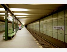 U-Bahn-Station mit grünlichen Wandfliesen, einer Sitzbank und einem Bahnsteig, der sich parallel zu den Gleisen erstreckt. Ein Schild mit der Aufschrift "Neu-Westend" ist an der Wand sichtbar, und im Hintergrund sind beleuchtete Werbung sowie der Tunnel zu erkennen.