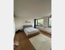 Schlafzimmer mit Doppelbett und hellem Bettbezug, hellem Holzboden und einem großen Läufer. An einer Wand befindet sich ein kleines Wandregal mit Dekorationsgegenständen, während große Fenster mit Blick ins Grüne auf der gegenüberliegenden Seite viel Licht hereinlassen.