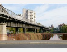 Eine Straßenbrücke aus Metall führt über einen Kanal, umgeben von grüner Vegetation. Im Hintergrund sind ein modernes Hochhaus und eine Kirche mit großer Kuppel sichtbar.